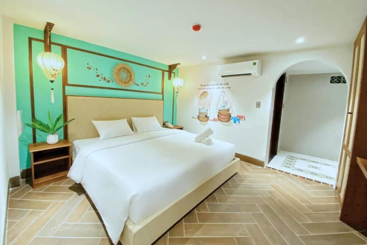 A hotel room with a large bed, a small bedside table, a potted plant, and a wall-mounted air conditioning unit.