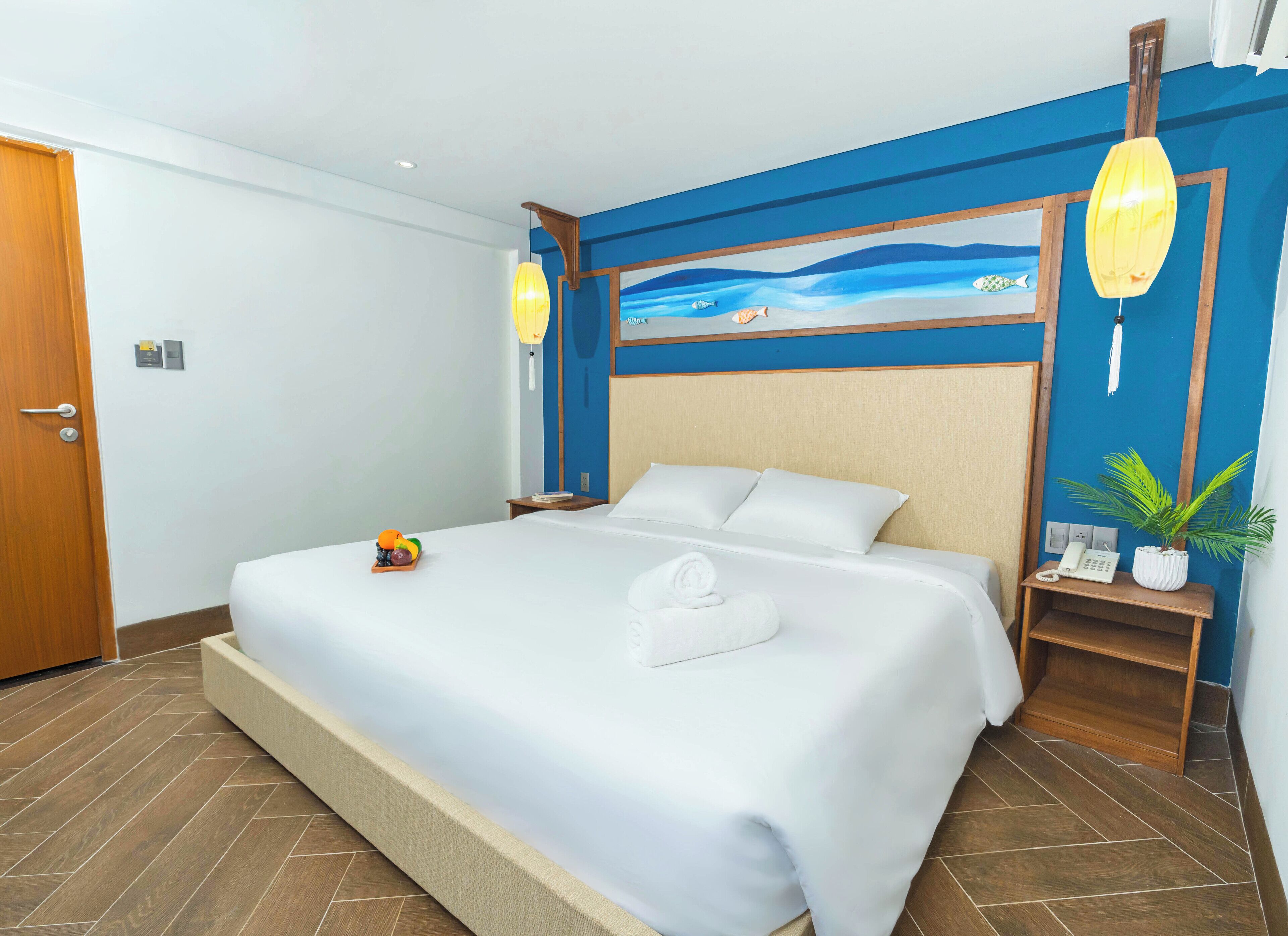 Una habitación de hotel con una cama grande, un cuadro de una playa en la pared, una mesita de noche de madera con un teléfono y una planta en maceta.
