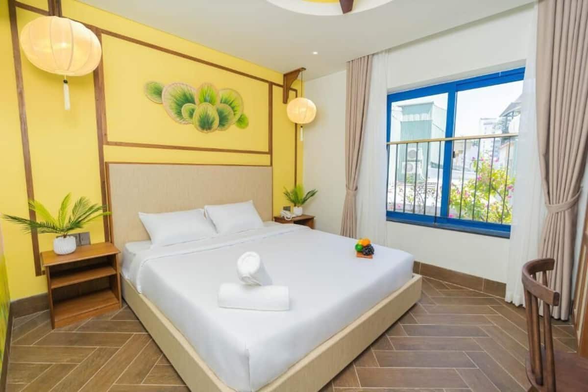 A hotel room with a large bed, a window with a view, a wall-mounted lamp, and a small table with a plant.