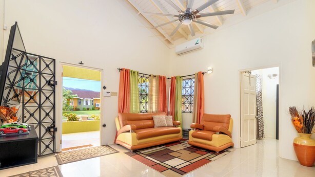 Uma sala de estar ampla com pé direito alto, ventilador de teto, sofá, duas poltronas, mesa de centro e um vaso decorativo.