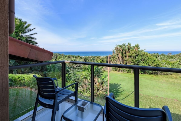 Un balcon avec deux chaises et une table, offrant une vue sur un paysage verdoyant et luxuriant ainsi que sur l’océan.