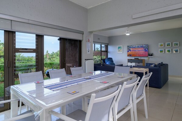 Un espace repas moderne avec une grande table, des chaises blanches et une vue sur la verdure environnante.