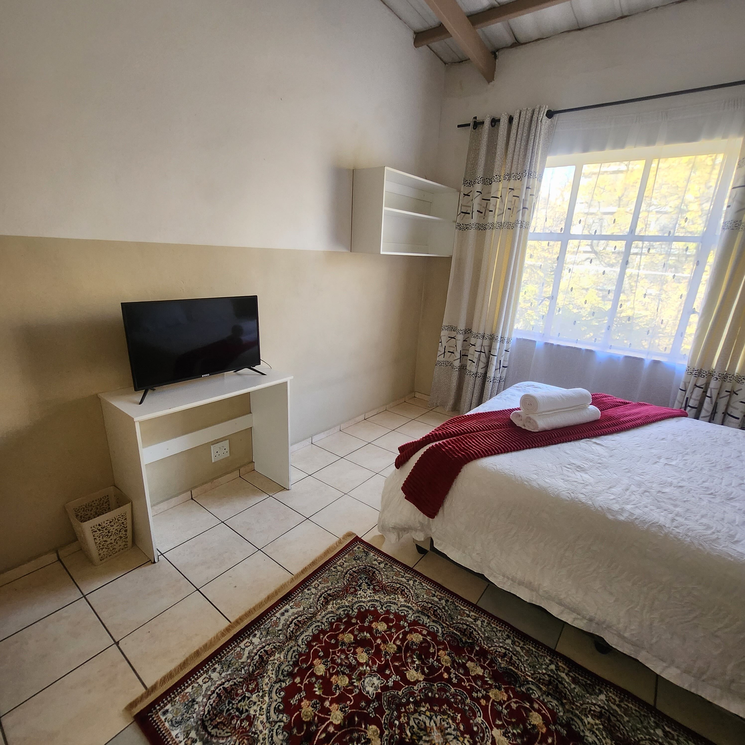 Une chambre à coucher avec un lit, un téléviseur posé sur un meuble, une fenêtre avec des rideaux et un tapis à motifs.