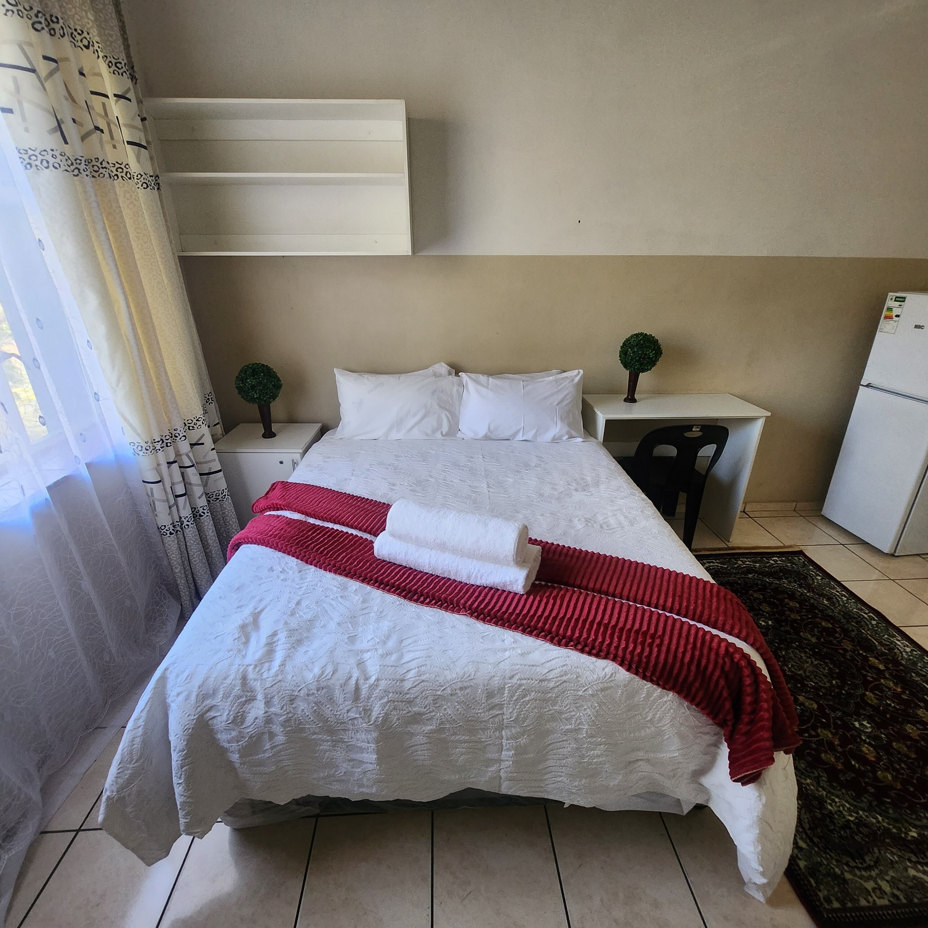 A neatly made bed with white and red linens, a small nightstand with a plant, and a refrigerator in a hotel room.