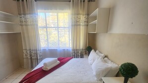 Een opgemaakt bed met witte lakens en een rode deken, een klein tafeltje met een vaas met groen, en een raam met gordijnen.