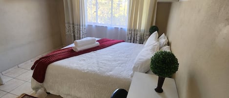 Ein Schlafzimmer mit einem Bett, einem Schreibtisch, einem Stuhl, einem Fenster mit Vorhängen und einer kleinen Topfpflanze.