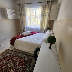 Une chambre à coucher avec un lit, un bureau, une chaise, une fenêtre avec des rideaux et une petite plante en pot.