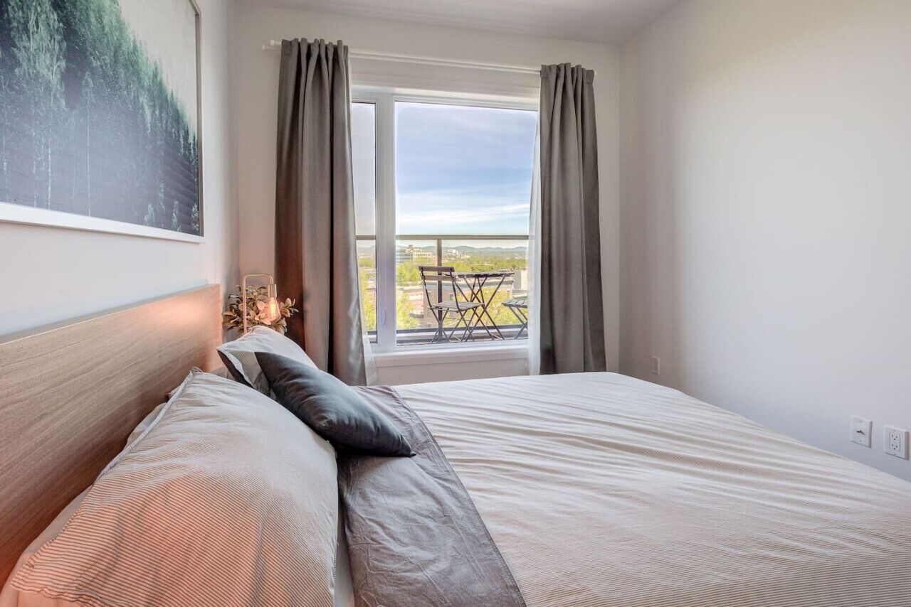 A bedroom with a large bed, a window with curtains, a balcony with a table and chairs, and a painting on the wall.
