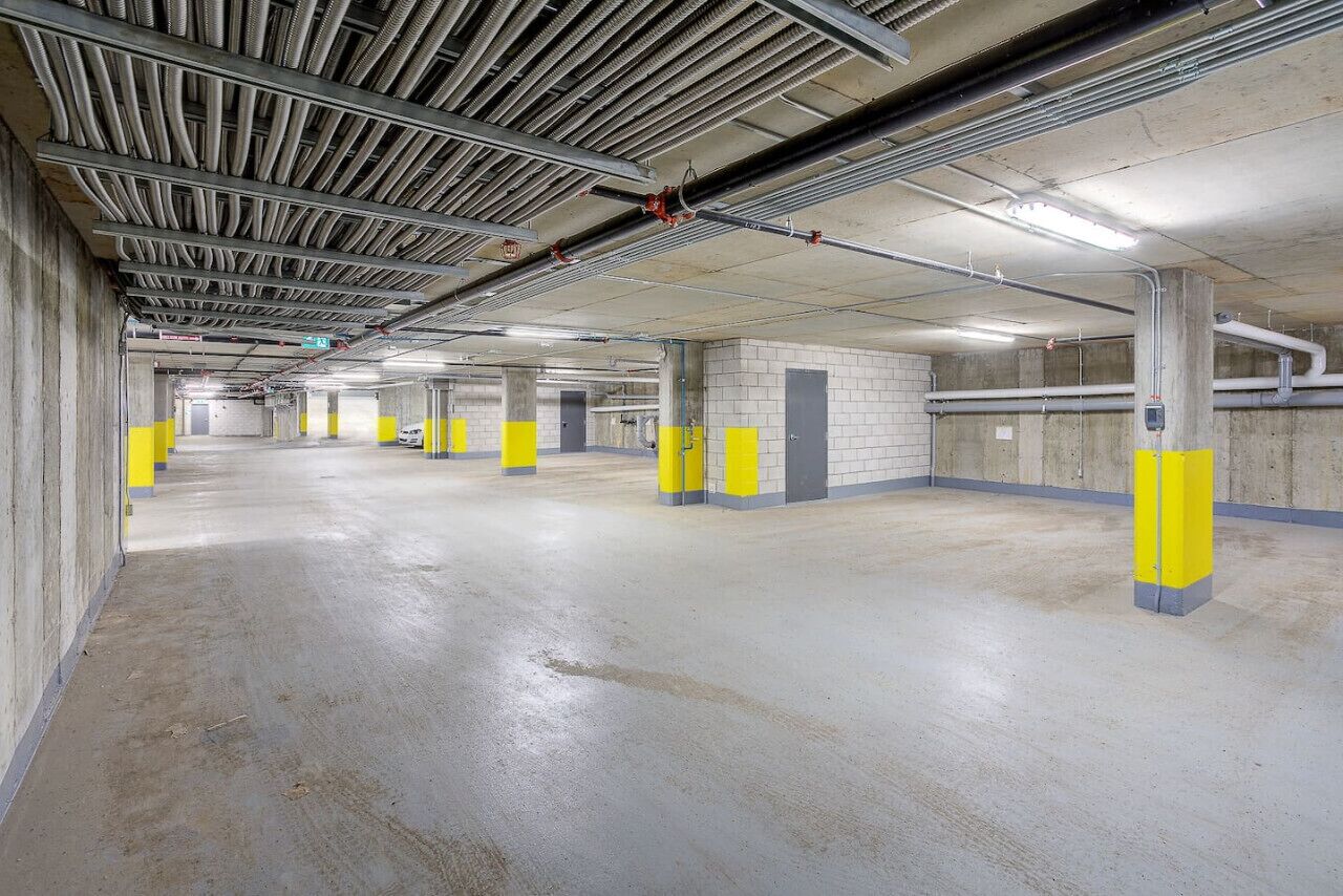 A multi-level parking garage with concrete floors and walls, yellow safety posts, and visible structural beams and pipes.