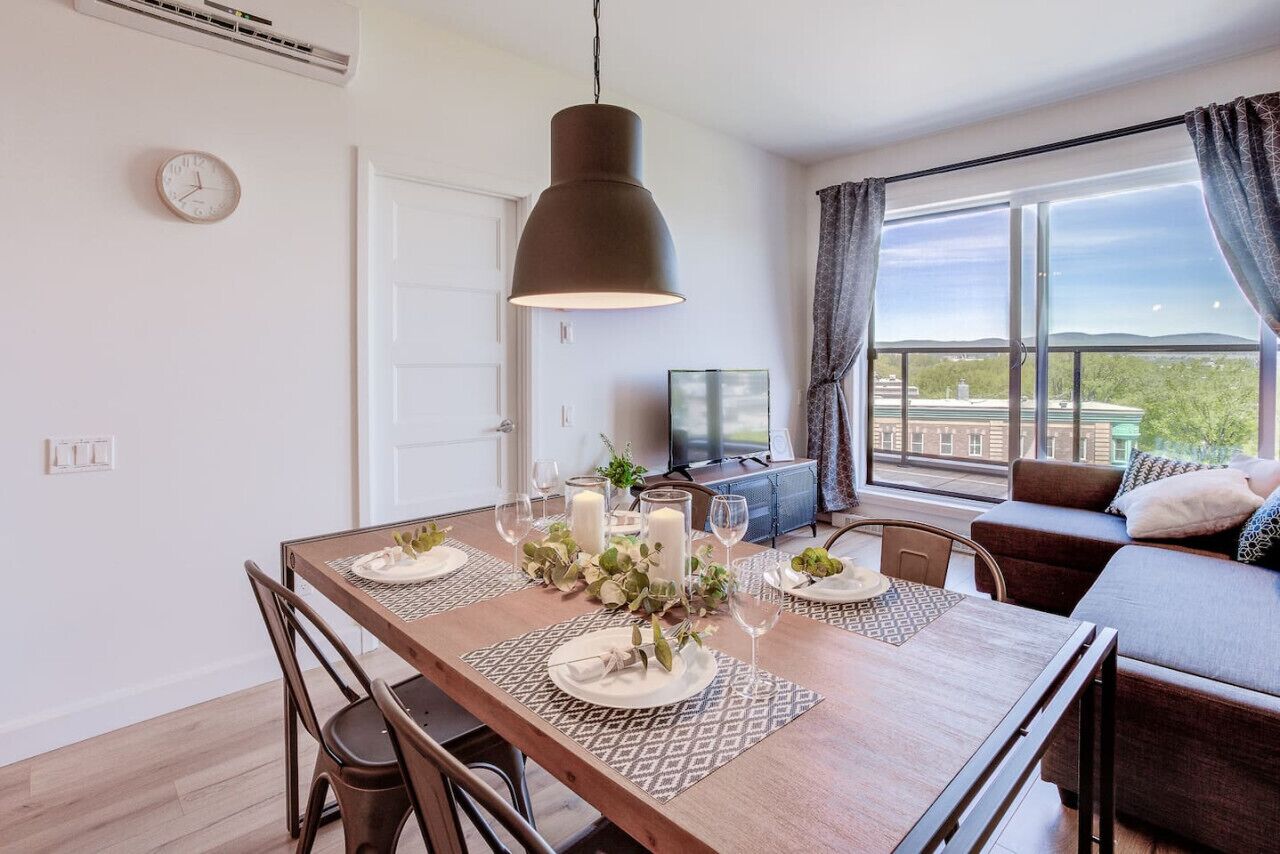 A dining area with a table set for four, a hanging pendant light, a sofa, and a large window with a view.