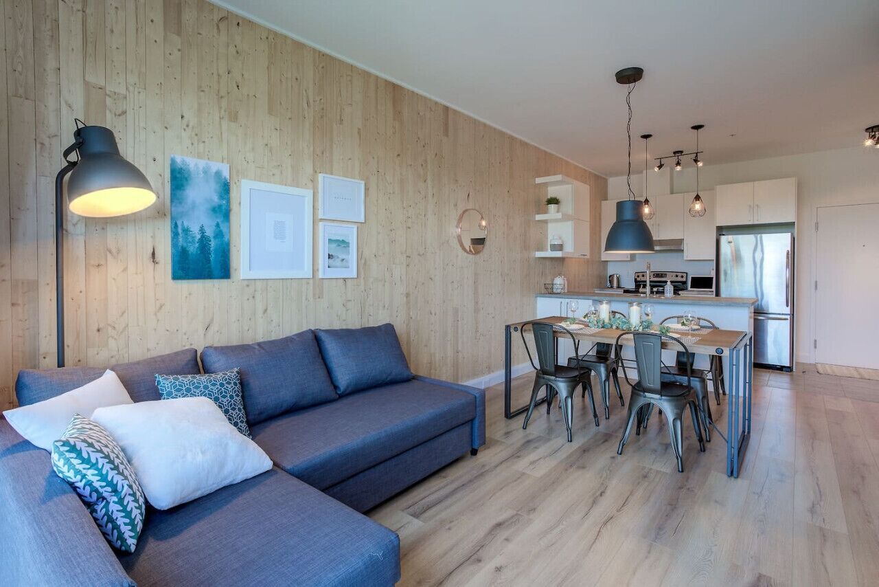 A modern living room with a sofa, a dining table with chairs, a kitchen area, and a wooden wall.