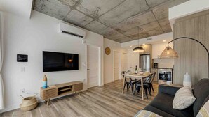 A modern living room with a flat-screen TV, wooden flooring, a sofa, and a dining area with chairs and a table. - Le Jules-Rooftop Pool (Québec)