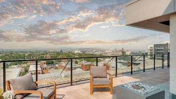 A rooftop terrace with two wooden chairs and a small planter, overlooking a cityscape with buildings and a distant smokestack.