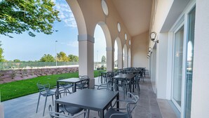 An outdoor patio with multiple tables and chairs, a stone wall, and a view of a tennis court. - I Campi Wine Relais (Lazise)