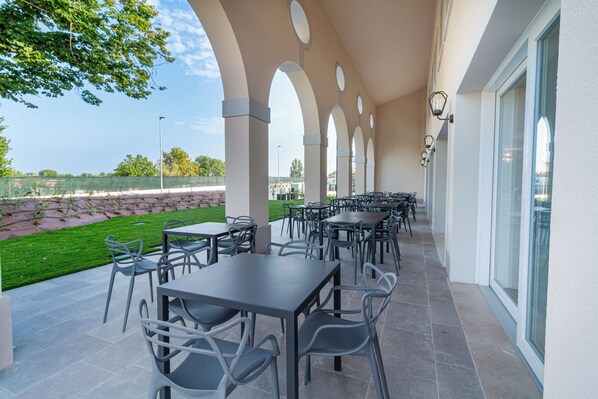 An outdoor patio with multiple tables and chairs, a stone wall, and a view of a tennis court.