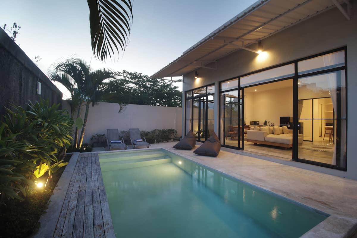Een modern huis met een zwembad, een terras met lounge stoelen en een glazen deur die leidt naar een woonruimte.