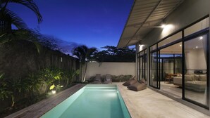 Un área de piscina moderna al aire libre con cubierta de hormigón, sillones de descanso y un espacio habitable con paredes de vidrio.