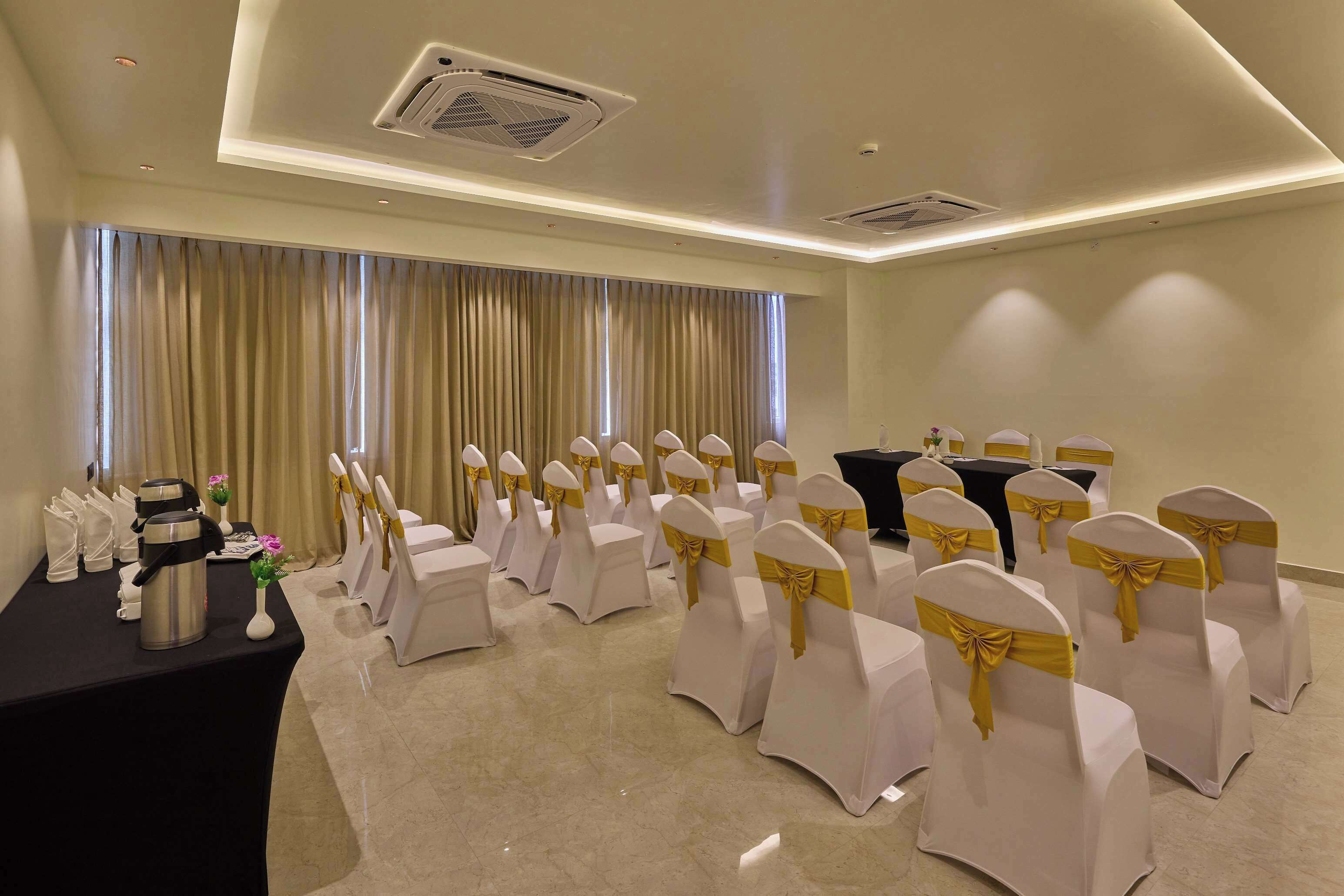 A conference room with rows of white chairs with gold sashes, a black table with a coffee pot and a vase with flowers, and a large window with curtains.