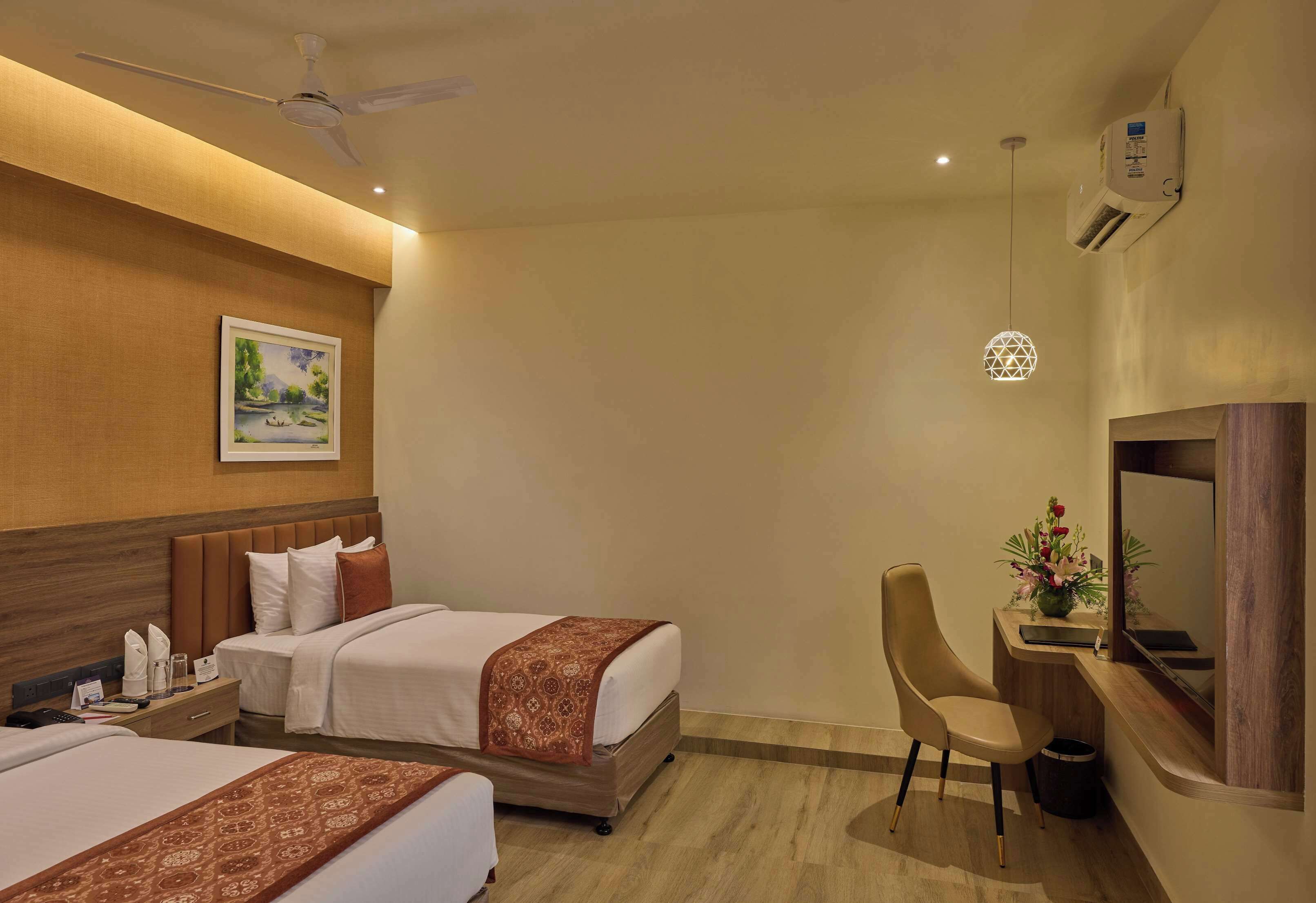 A hotel room with two beds, a desk with a chair, a TV, a ceiling fan, and an air conditioning unit.