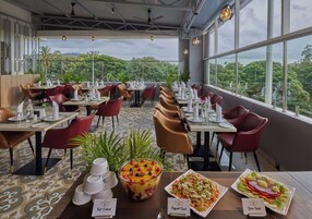 Un restaurant avec des tables dressées pour le service, une grande fenêtre donnant sur un paysage verdoyant et un ventilateur de plafond.