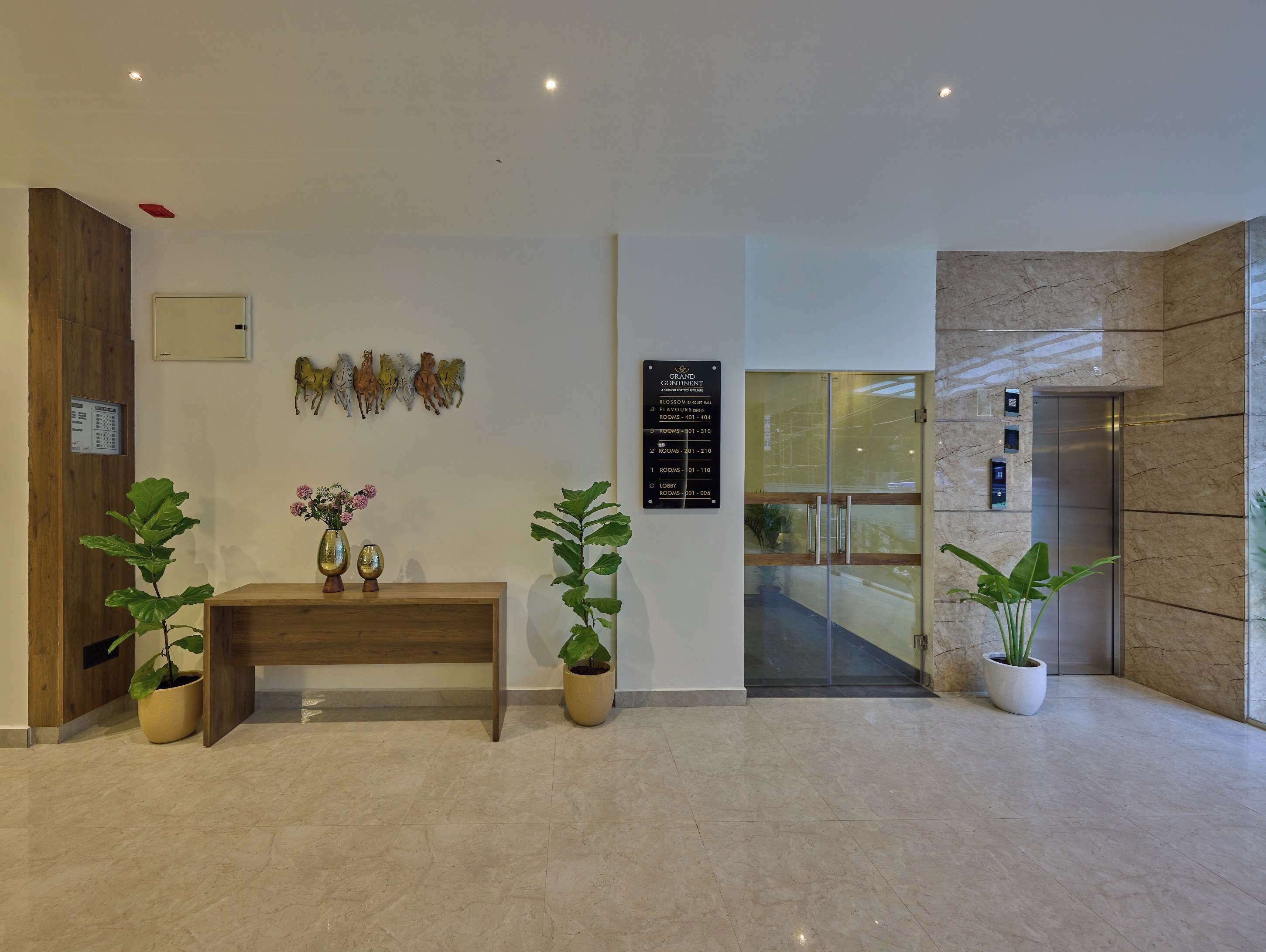 Un vestíbulo de hotel con mostrador de recepción, plantas en macetas, un letrero en la pared con los números de las habitaciones y un ascensor.