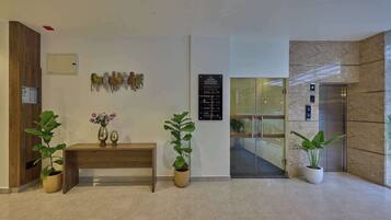 Un vestíbulo de hotel con mostrador de recepción, plantas en macetas, un letrero en la pared con los números de las habitaciones y un ascensor.