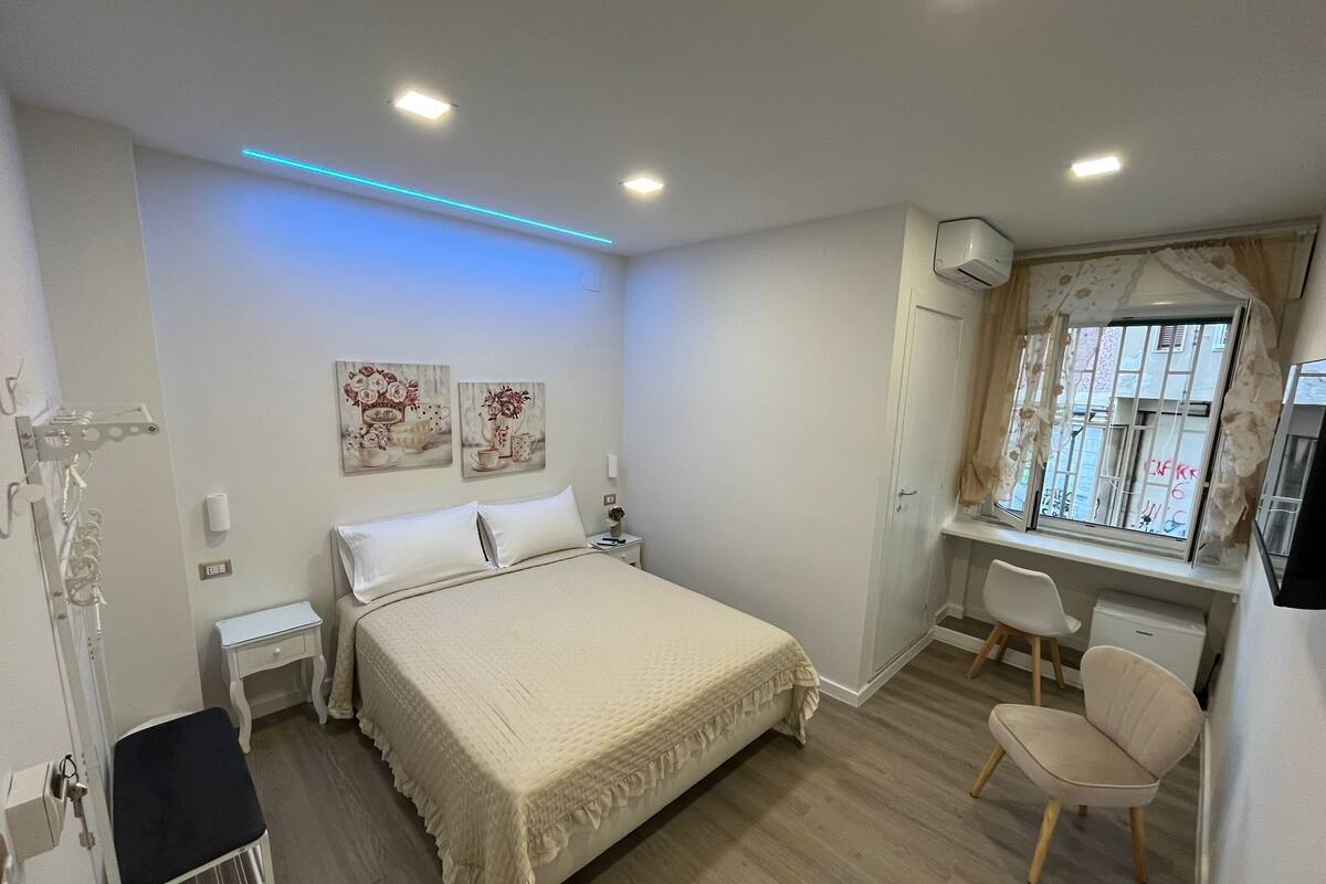 A bedroom with a bed, bedside tables, a desk with a chair, a window with curtains, and wall art.