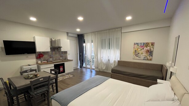 A modern hotel room with a large bed, a dining area with a table and chairs, a kitchenette, a flat-screen TV mounted on the wall, and a window with curtains. - F.ROOMS NAPOLI VIA SEMENTINI (Naples)