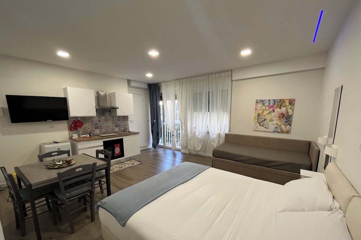A modern hotel room with a large bed, a dining area with a table and chairs, a kitchenette, a flat-screen TV mounted on the wall, and a window with curtains.
