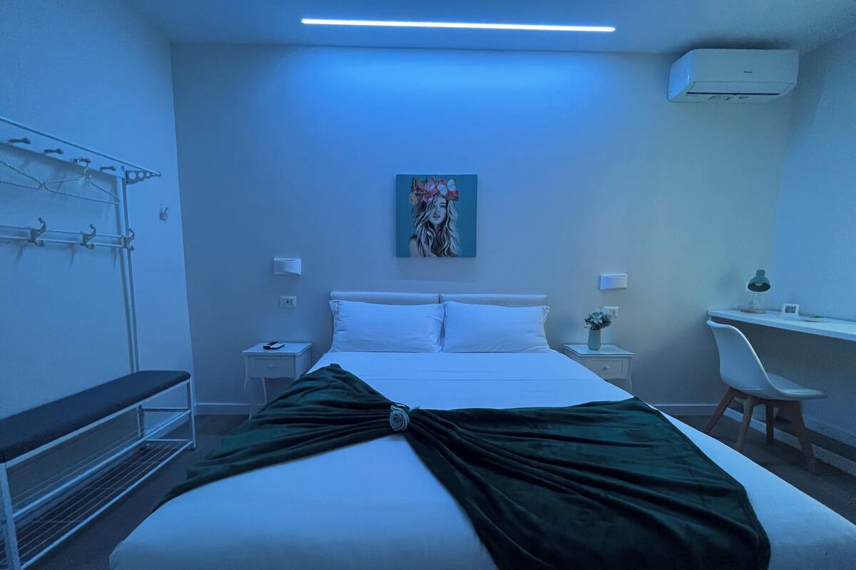 A hotel room with a bed, bedside tables, a desk, a chair, a wall-mounted air conditioner, and a painting.