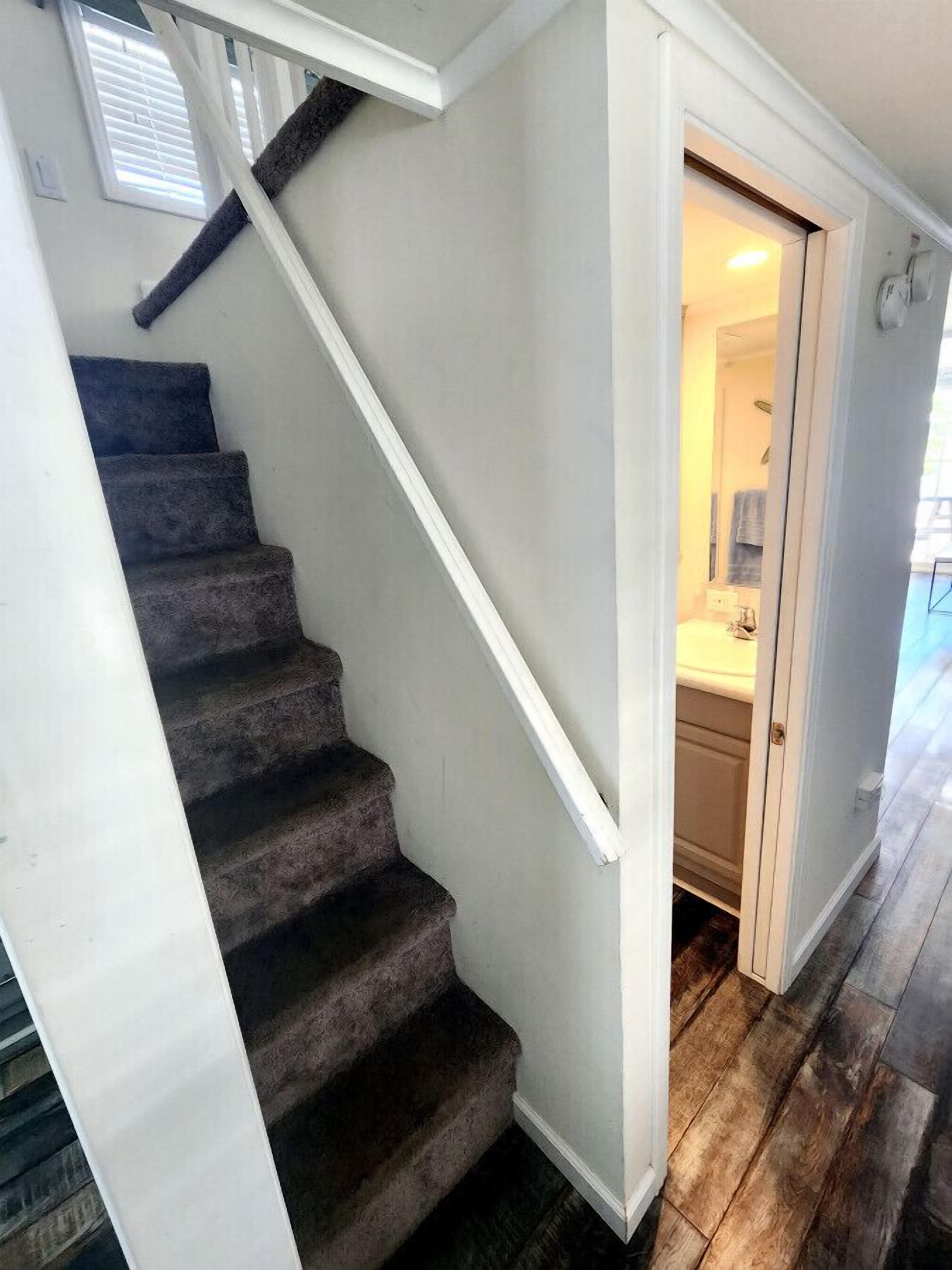 Een trap met donkere, beklede treden en witte afwerking, die leidt naar een badkamer met een witte wastafel en spiegel.