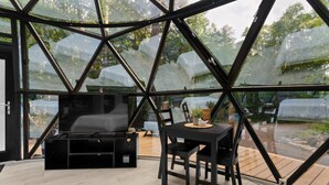 A modern interior with a glass enclosure, a black sofa, a dining table with chairs, and a television. - Allure Muskoka Glass Dome (Huntsville)