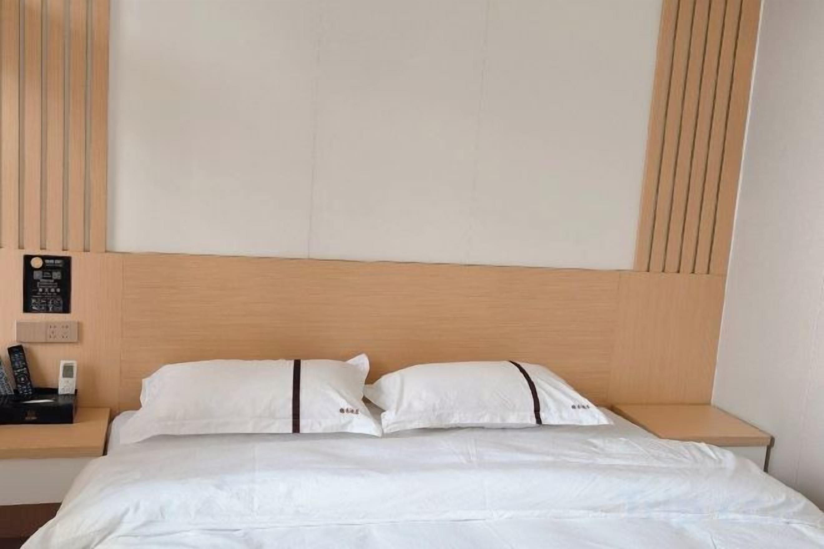 A hotel room with a wooden headboard, two white pillows, a bedside table with a phone, and a wall-mounted control panel.