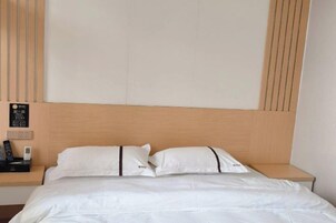 Quarto de hotel com cabeceira de madeira, duas almofadas brancas, mesa de cabeceira com telefone e painel de controle fixado na parede.