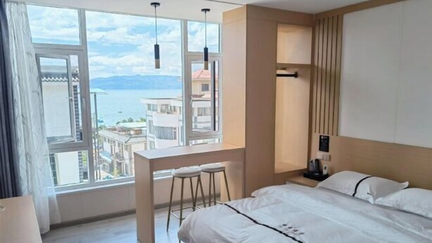 A modern hotel room with a large window offering a view of the sea and buildings. There is a bed with white linens, a small desk with two stools, and a nightstand with a lamp.