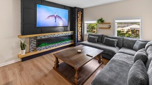 Een woonkamer met een grote flatscreen televisie, een open haard, een hoekbank en een salontafel.
