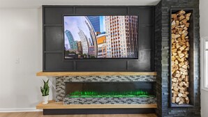 Een moderne woonkamer met een flatscreen televisie boven een open haard, een stapel hout aan de rechterkant en een potplant aan de linkerkant.
