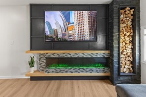 Ein modernes Wohnzimmer mit einem flachbildfernseher über einem Kamin, einem Holzstoß auf der rechten Seite und einer Topfpflanze auf der linken Seite.