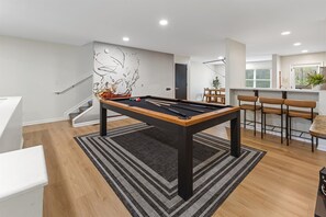 A pool table in a modern living room with a staircase, a dining area, and a kitchen in the background.