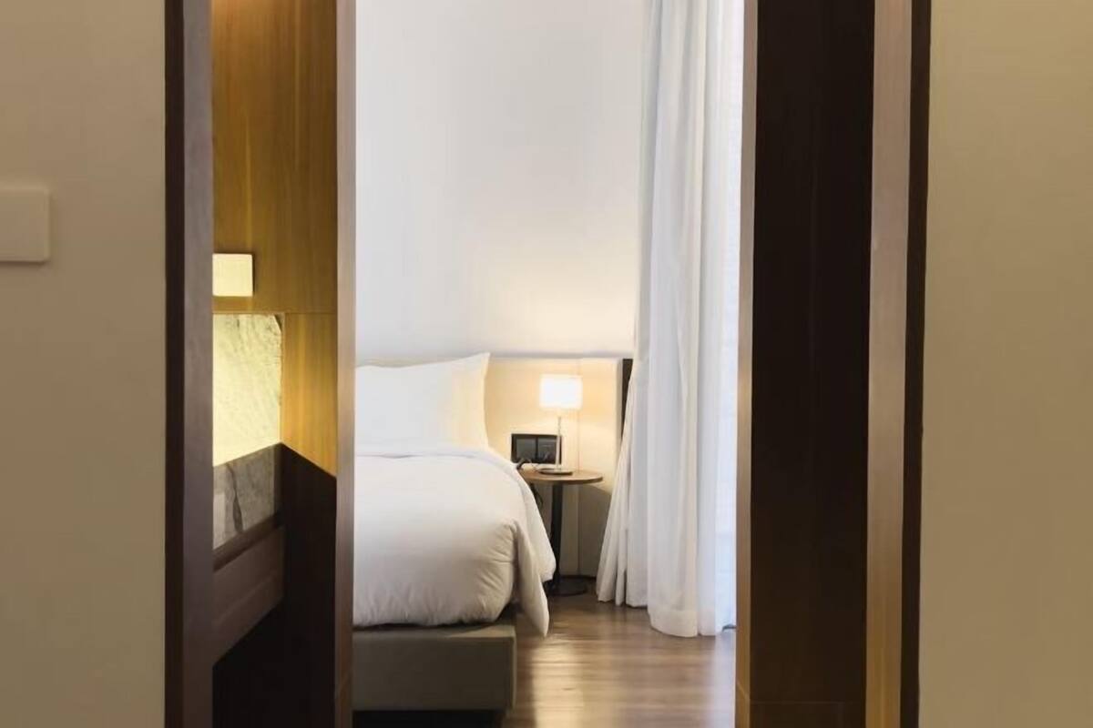 A hotel room with a bed, bedside table, lamp, and a view of the hallway.