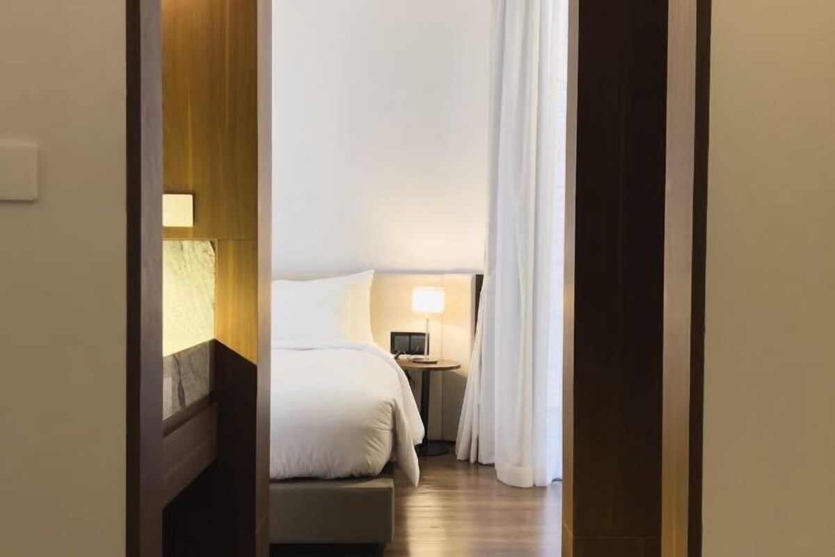 A hotel room with a bed, bedside table, lamp, and a view of the hallway.
