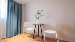 Una habitación con una mesa redonda, dos sillas blancas, un florero con planta y una cortina azul.
