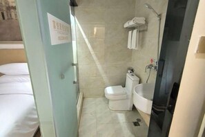 Un baño de hotel con inodoro, lavamanos y ducha. Hay un cartel en la puerta y se ve una cama en la habitación contigua.
