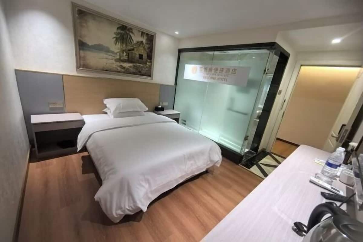 Habitación de hotel con una cama, una mesita de noche, un escritorio con una silla, un cuadro en la pared y una puerta de cristal con un letrero.