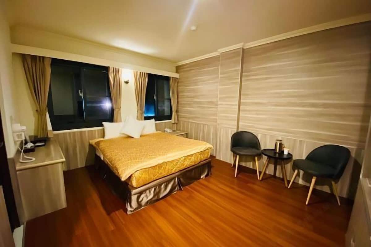 A hotel room with a bed, bedside table, lamp, two chairs, and a window with curtains.