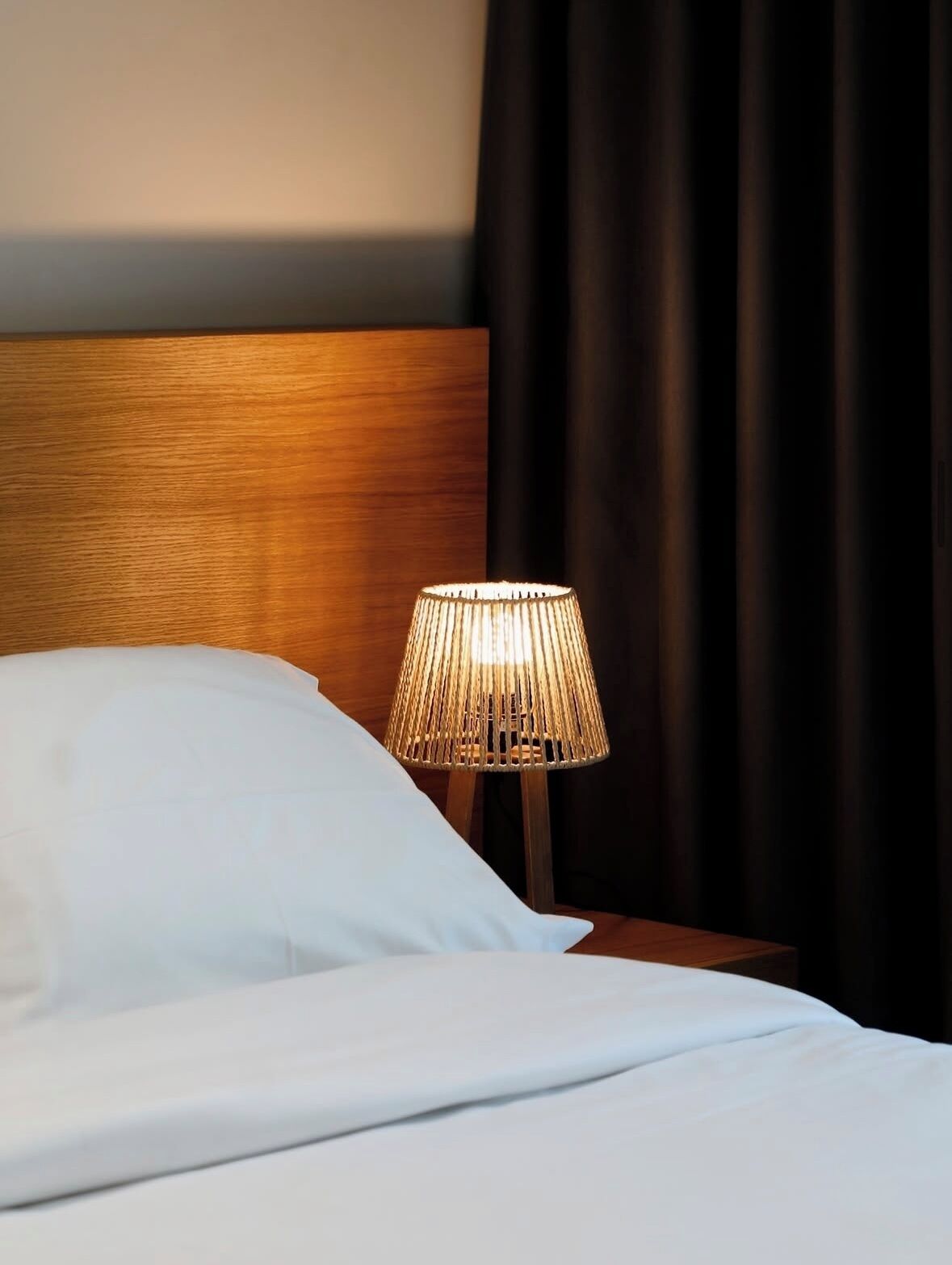 Una cama con ropa de cama blanca, cabecera de madera y lámpara con pantalla rayada.