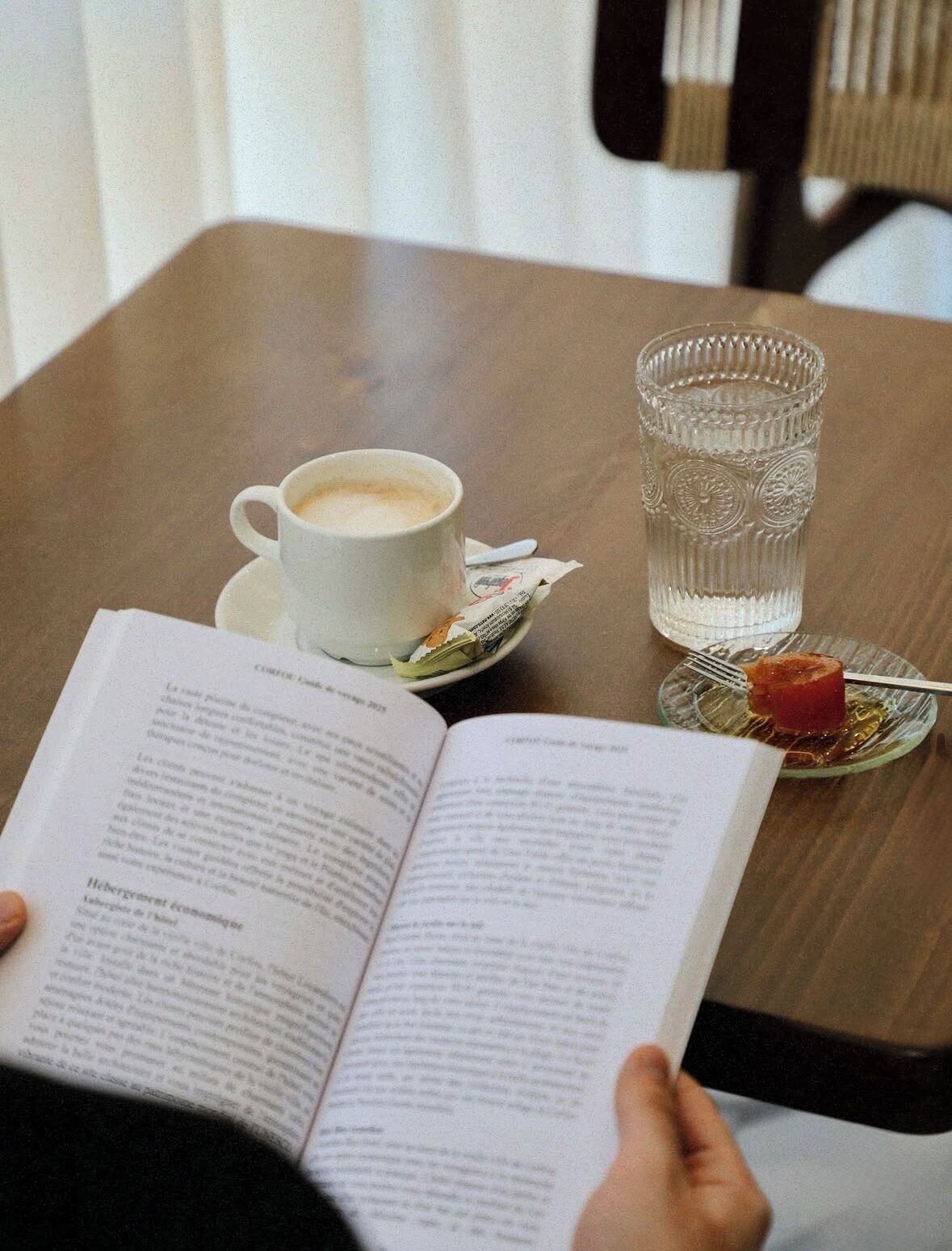 A person reading a book at a table with a cup of coffee, a glass of water, and a plate with a piece of food.