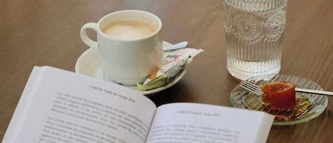 A person reading a book at a table with a cup of coffee, a glass of water, and a plate with a piece of food.