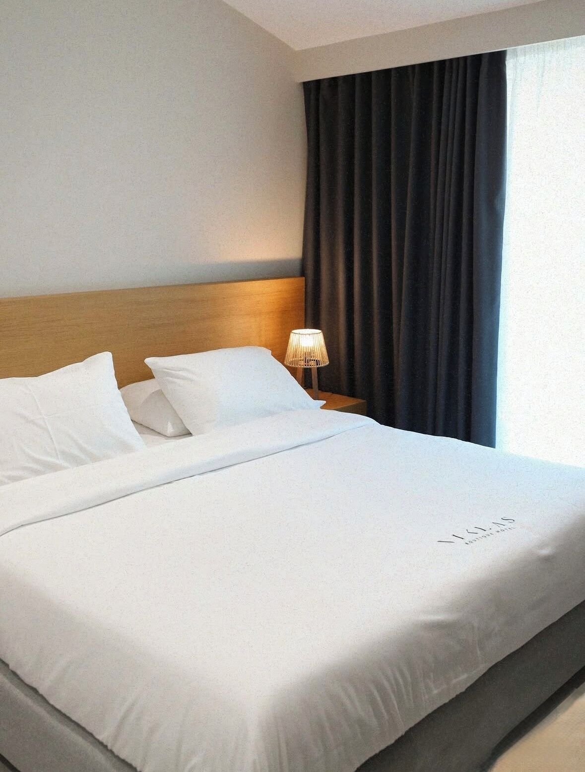 Habitación de hotel con una cama grande, cabecera de madera, lámpara de noche y ventana con cortinas oscuras.