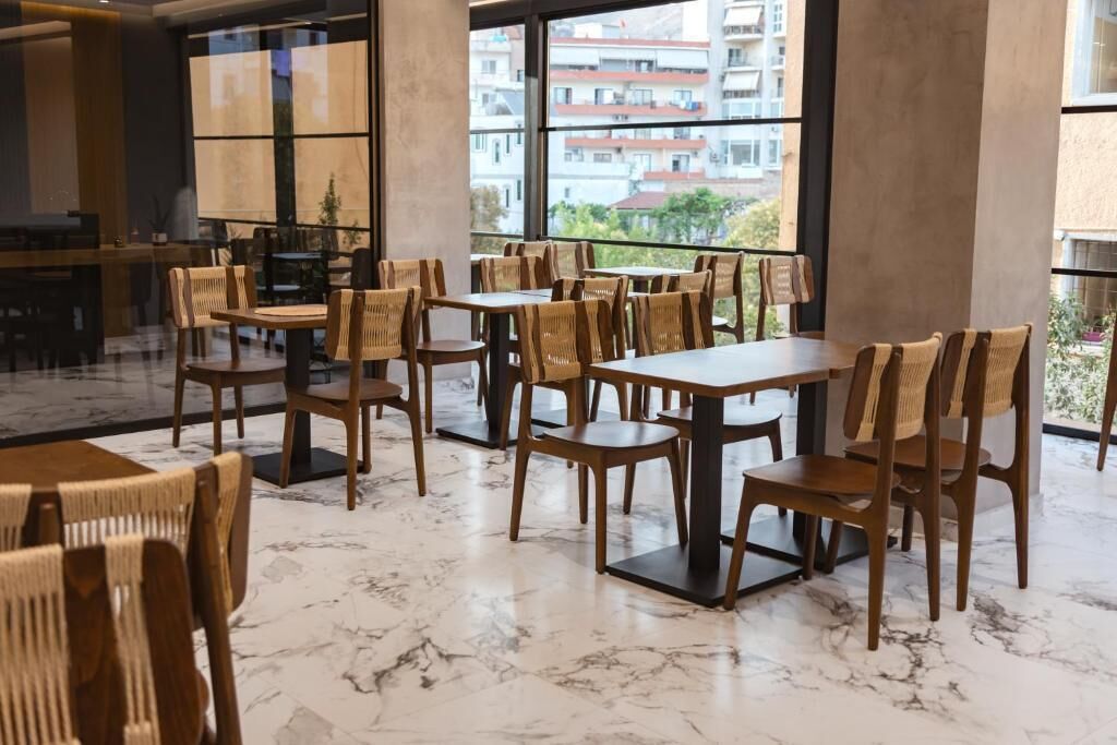 A spacious dining area with wooden tables and chairs, large windows offering a view of buildings outside.
